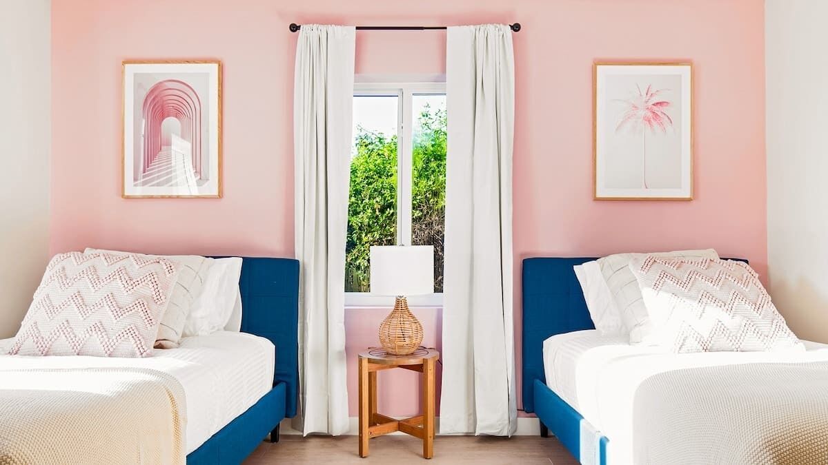 Een slaapkamer met twee bedden, een raam met witte gordijnen en ingelijste foto's aan de muur.