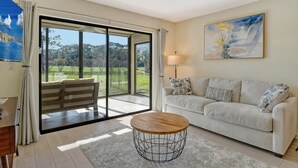 Uma sala de estar espaçosa com uma grande porta de vidro que se abre para um jardim, uma mesa de centro redonda e um sofá branco com almofadas estampadas.