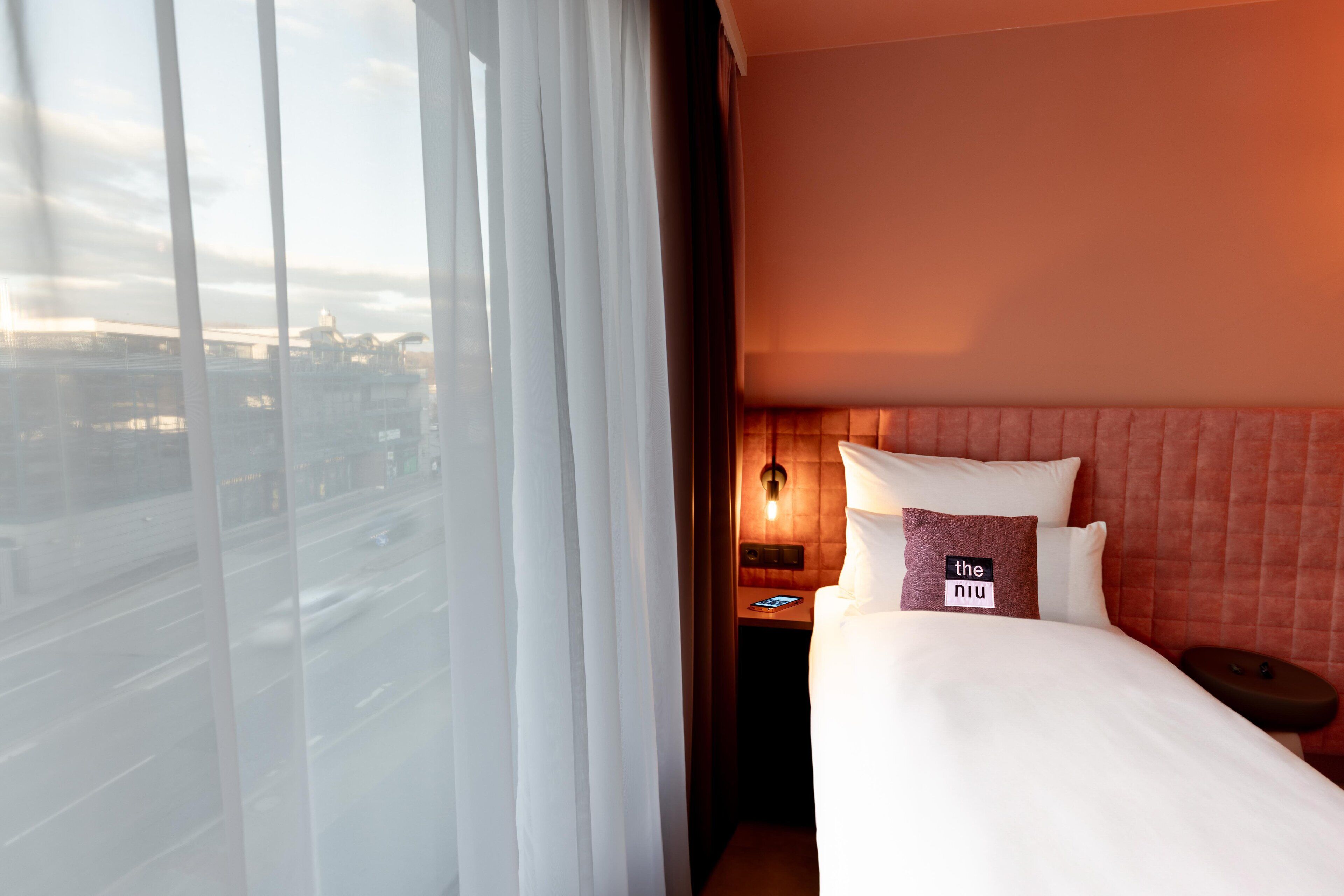 A hotel room with a large window, a bed with white bedding, a nightstand with a lamp, and a pillow with a label.