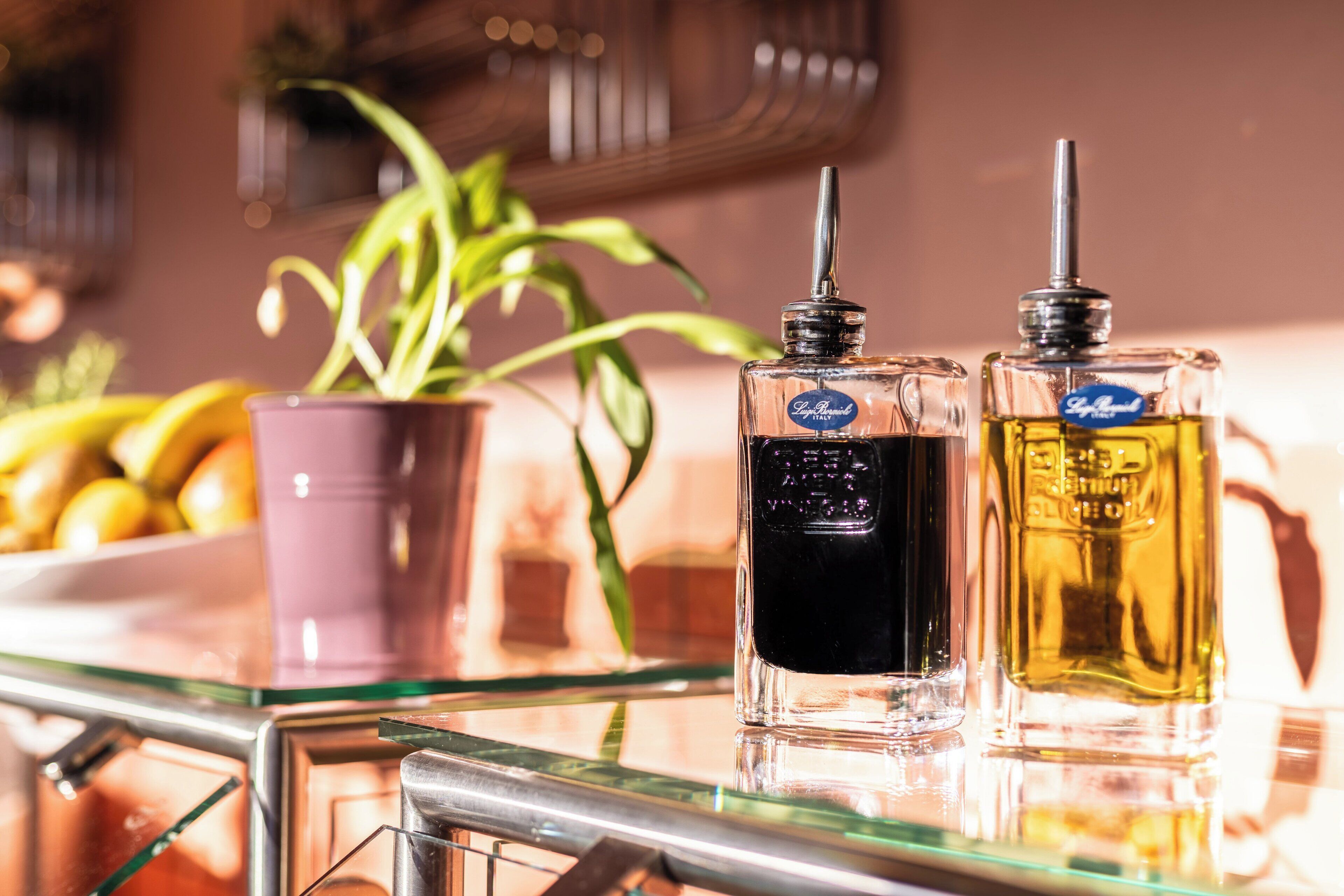 Dos botellas de perfume sobre una mesa de vidrio, con una planta en maceta y un frutero al fondo.