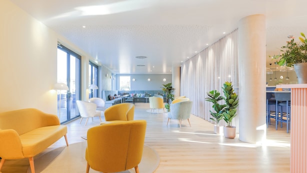 Eine geräumige, moderne Hotelhalle mit einer zentralen Sitzgruppe, bestehend aus gelben Sesseln, einem runden Teppich und Topfpflanzen.