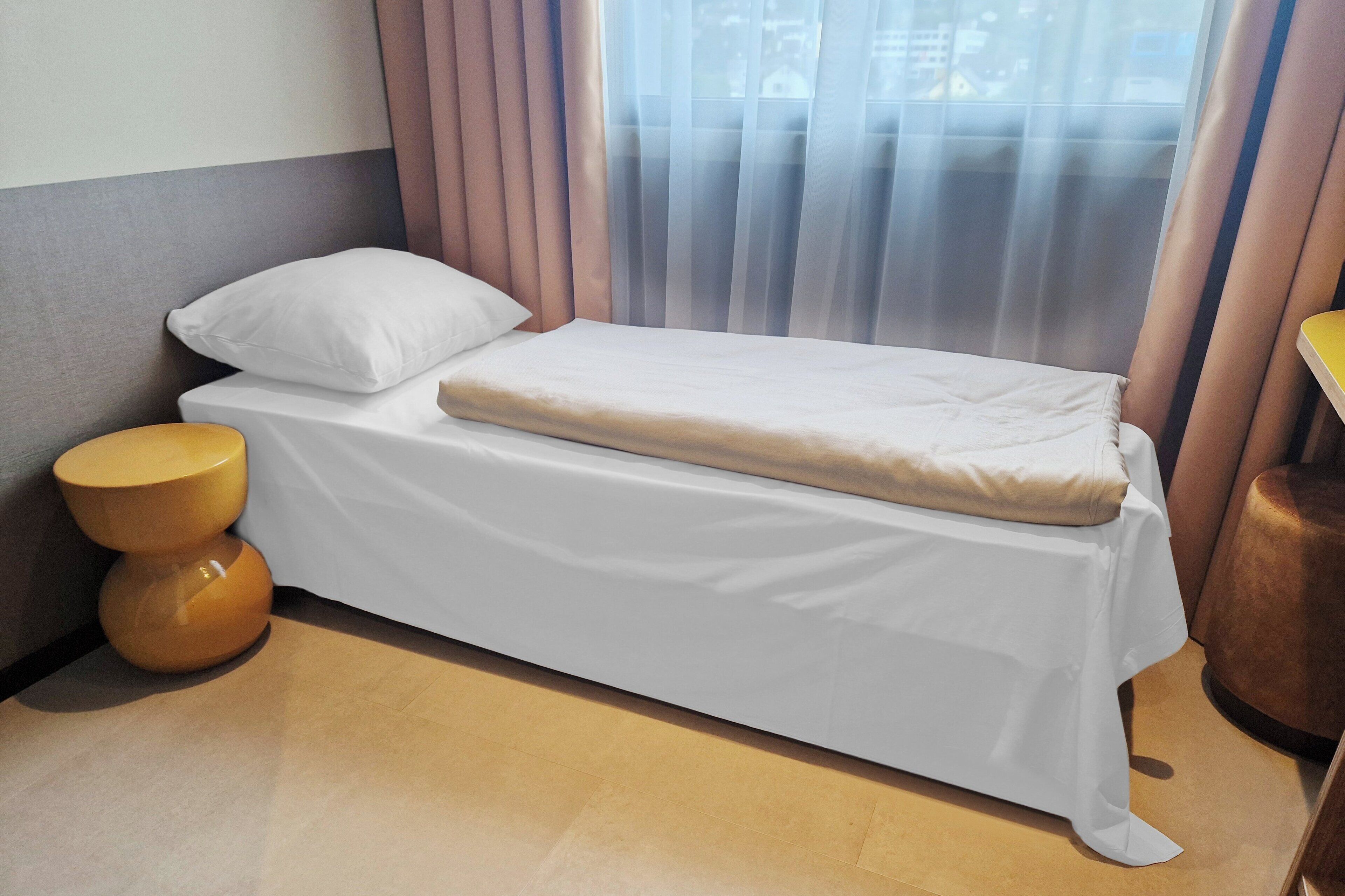 A single bed with a white mattress and pillow, a wooden nightstand, and a window with sheer curtains.
