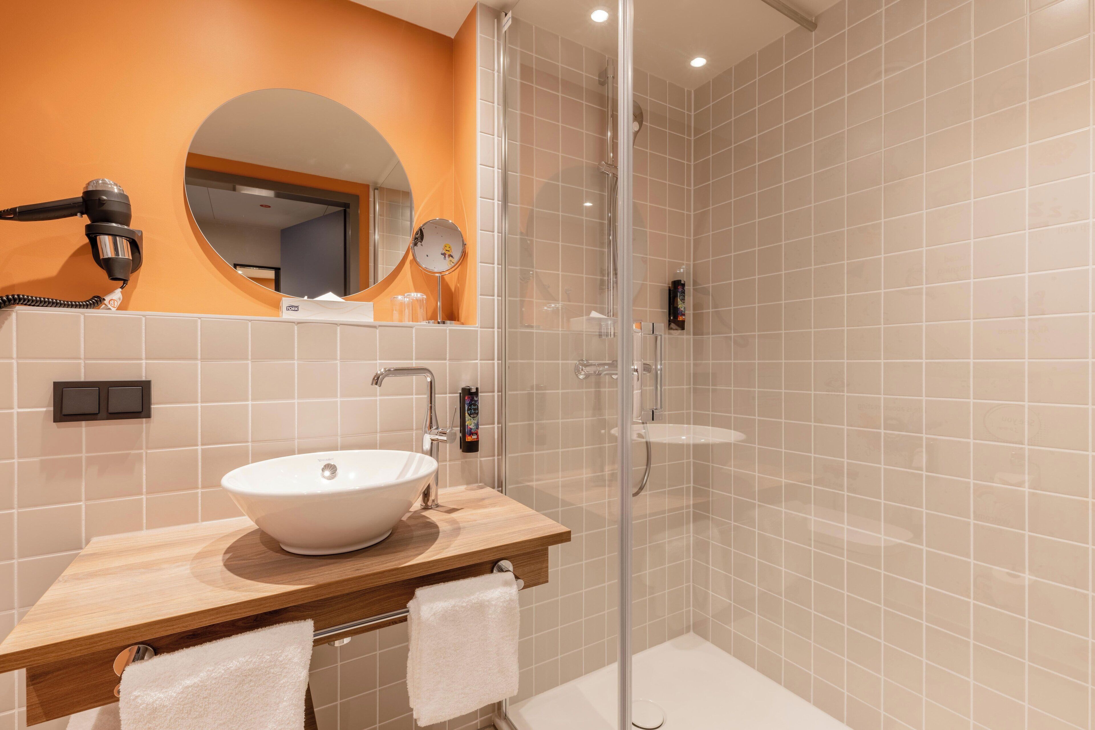 Un baño moderno con tocador de madera, un espejo ovalado grande, un lavabo blanco y una zona de ducha con mampara de vidrio.