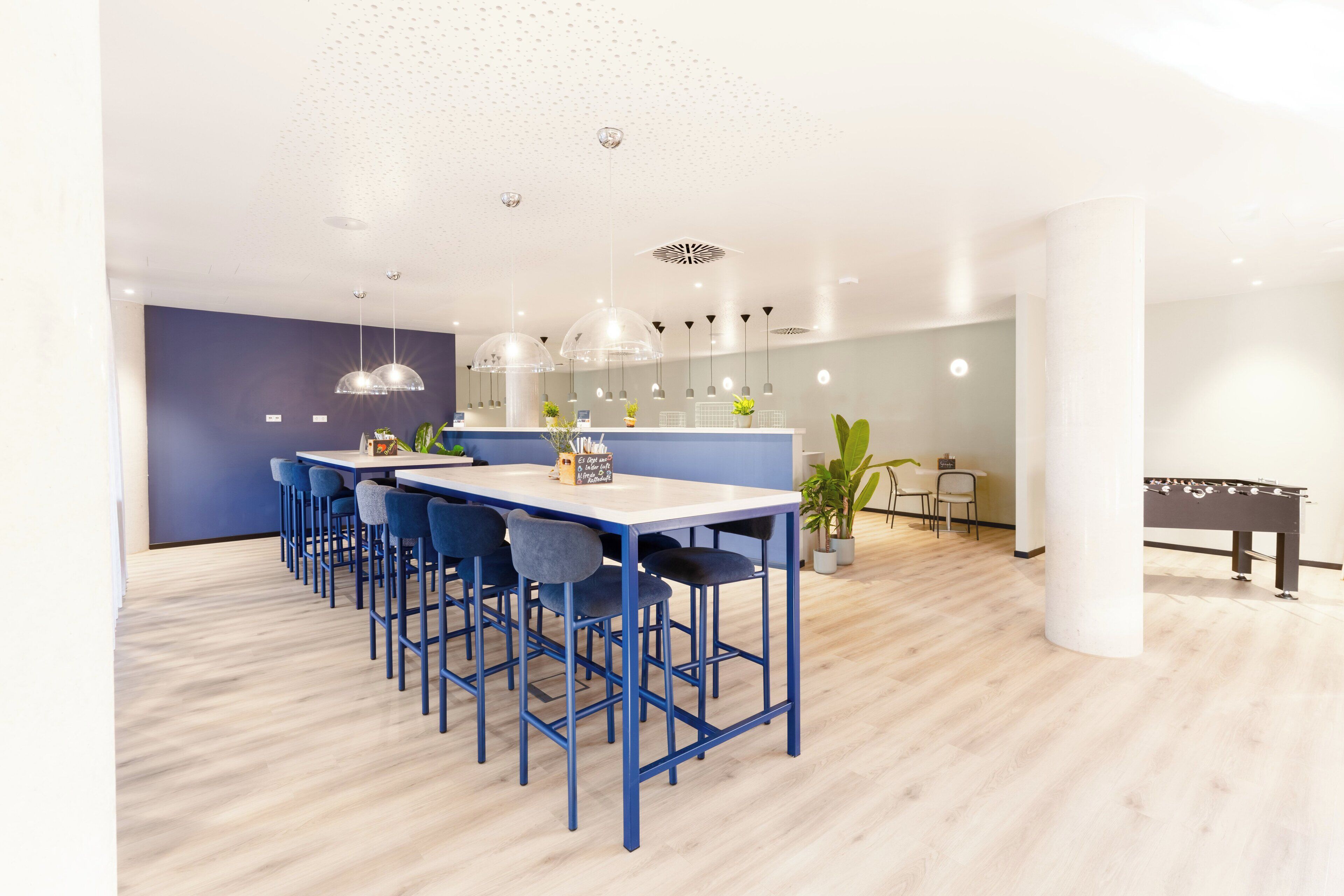 Un espace repas moderne avec une longue table et des chaises bleues, un coin bar avec des tabourets et une table de baby-foot.