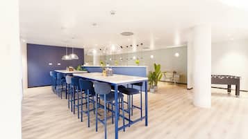 Un espace repas moderne avec une longue table et des chaises bleues, un coin bar avec des tabourets et une table de baby-foot.