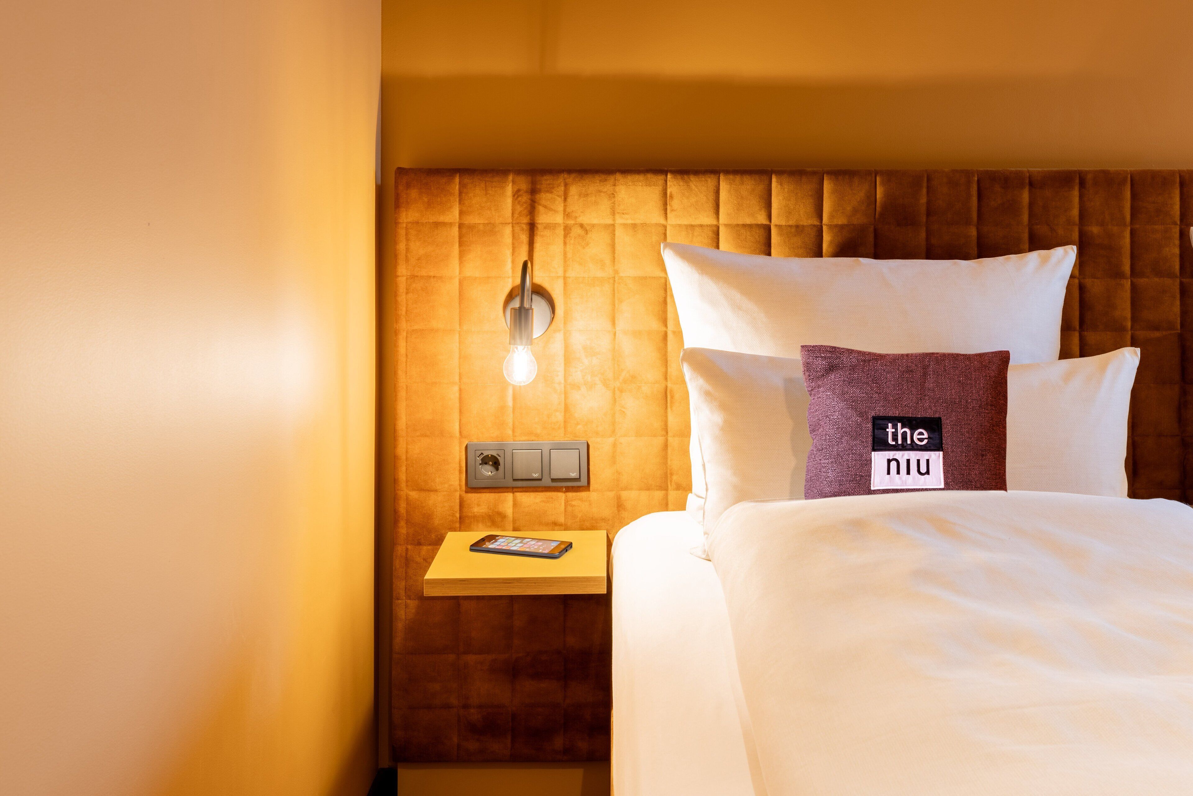A hotel room with a bed, a nightstand with a lamp, and a small table with a book.