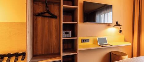 A hotel room with a wooden wardrobe, a mirror, a desk with a laptop, and a small round stool.