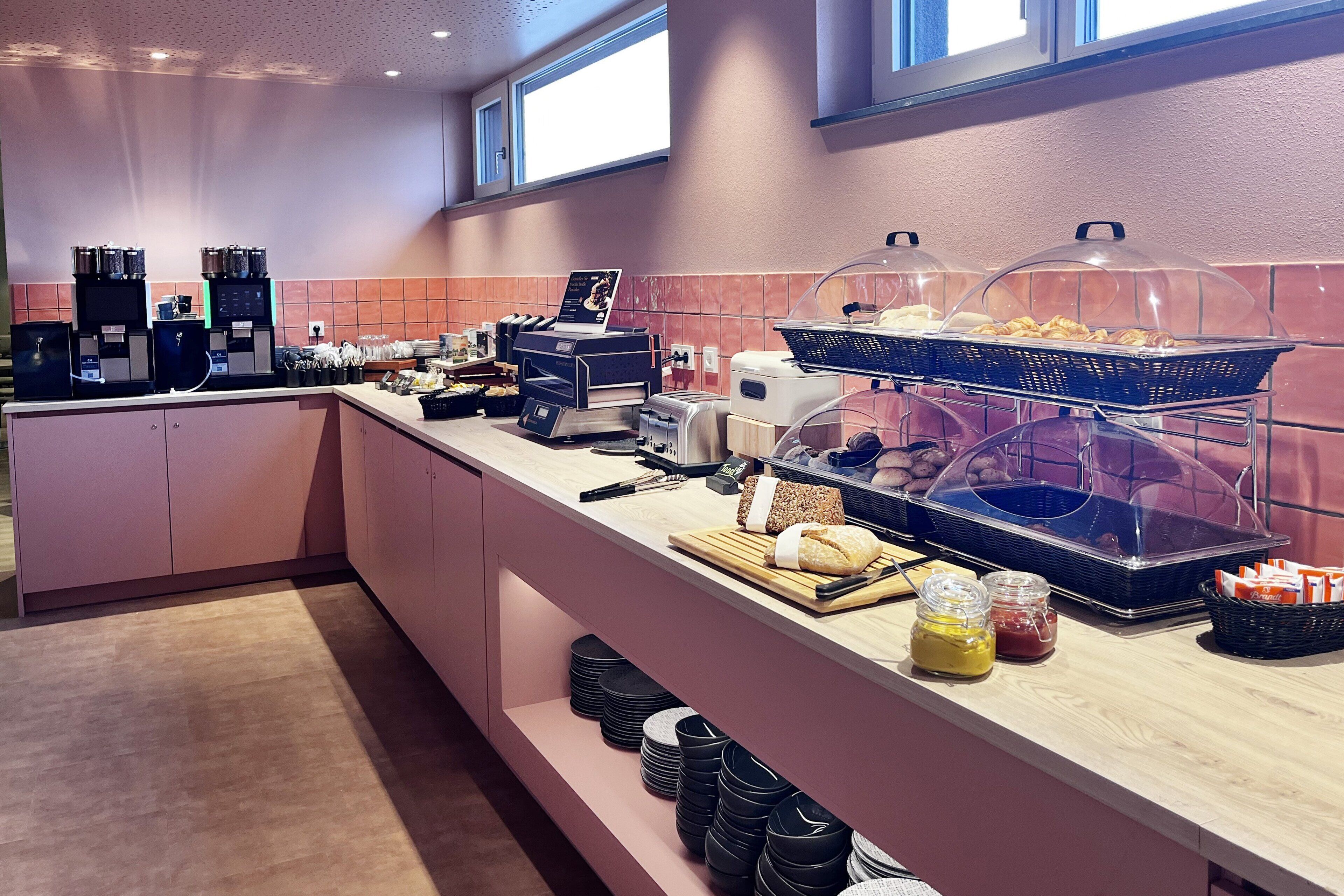 Zona de desayuno tipo buffet con máquinas expendedoras de café, diversos alimentos y acompañamientos.