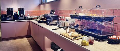 Zona de desayuno tipo buffet con máquinas expendedoras de café, diversos alimentos y acompañamientos.