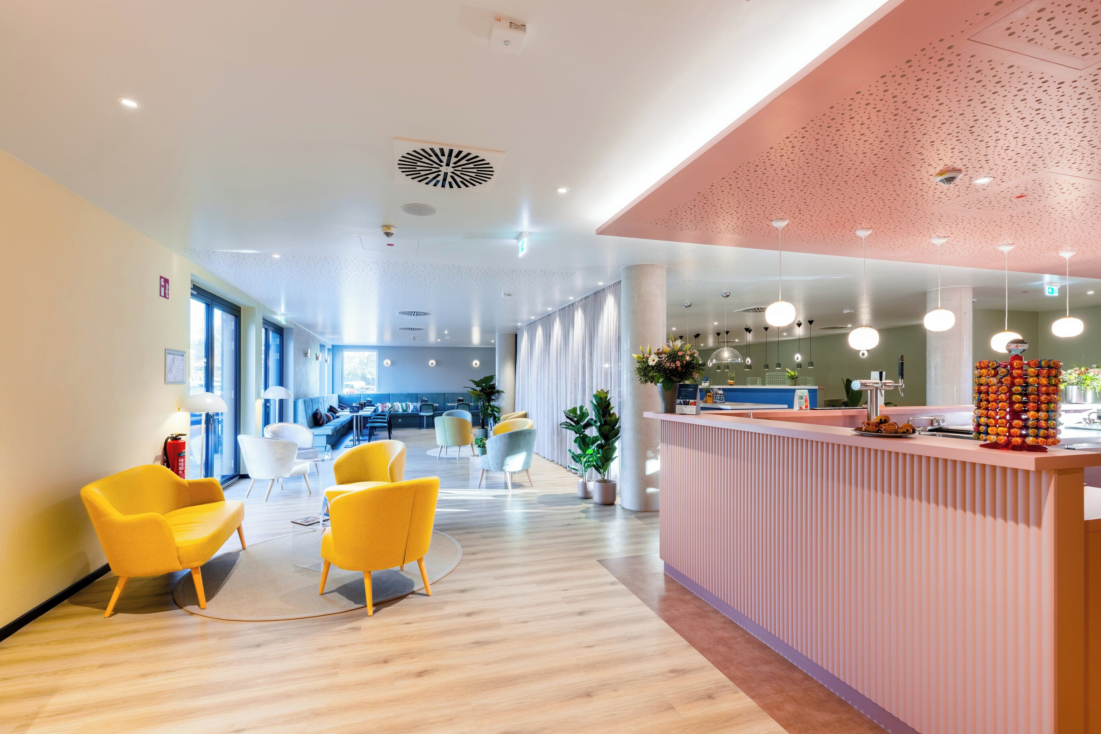 A modern reception area with a wooden floor, yellow chairs, a pink reception desk, and a large window providing natural light.