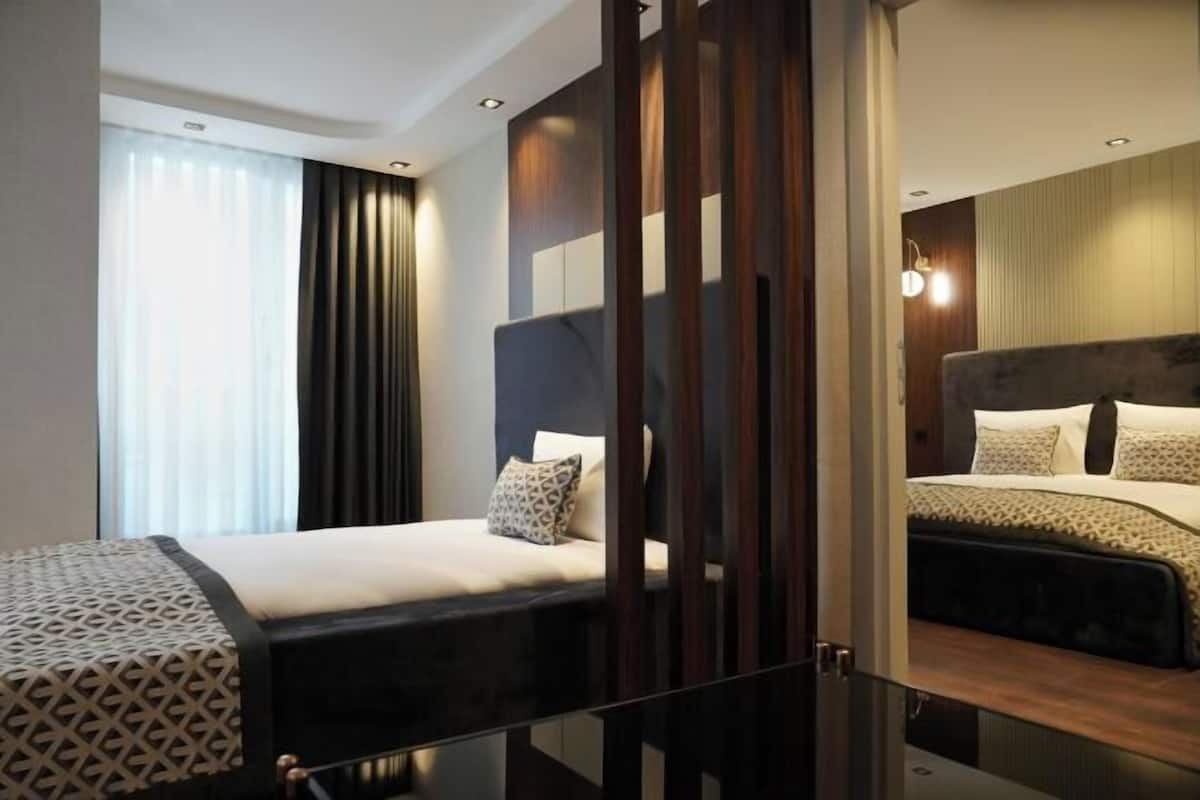 A hotel room with two beds, a large window with curtains, a wooden partition, and a modern light fixture.