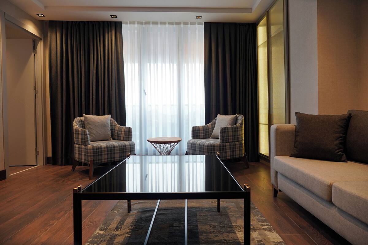 A modern living room with a glass coffee table, two plaid armchairs, a beige sofa, and a large window with sheer curtains.