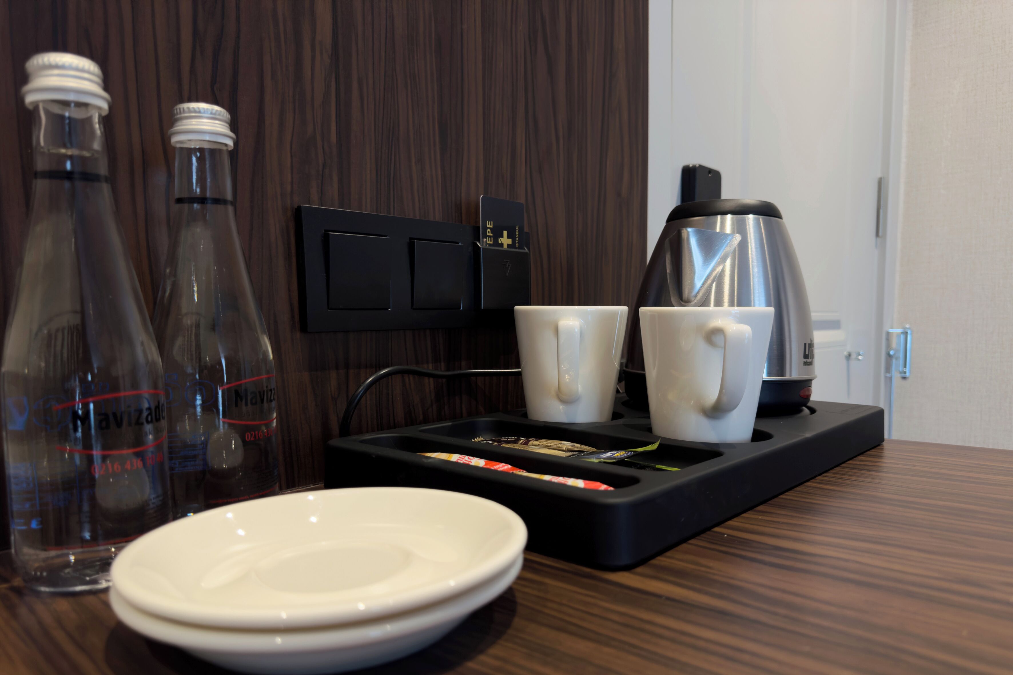 A hotel room with a wooden floor, a coffee maker, two cups, two bottles, and a tray with utensils.