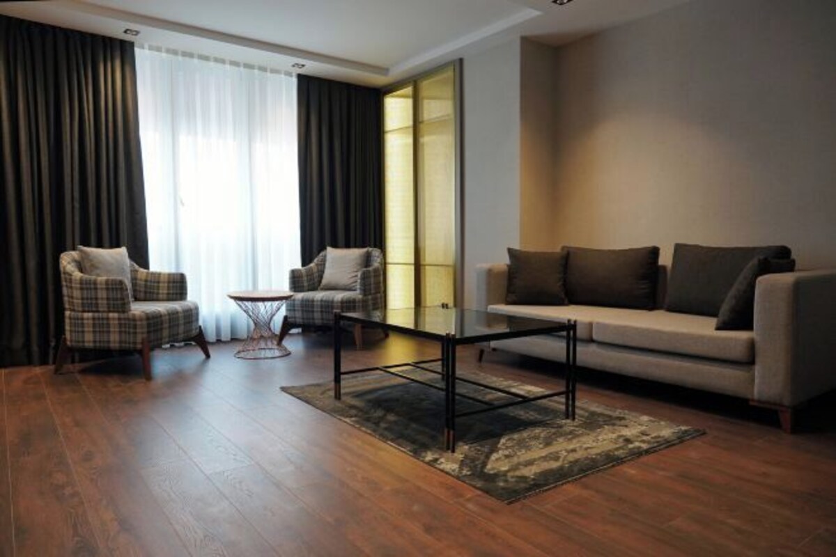 Una sala de estar moderna con un sofá, dos sillones, una mesa de centro de vidrio y un ventanal con cortinas.