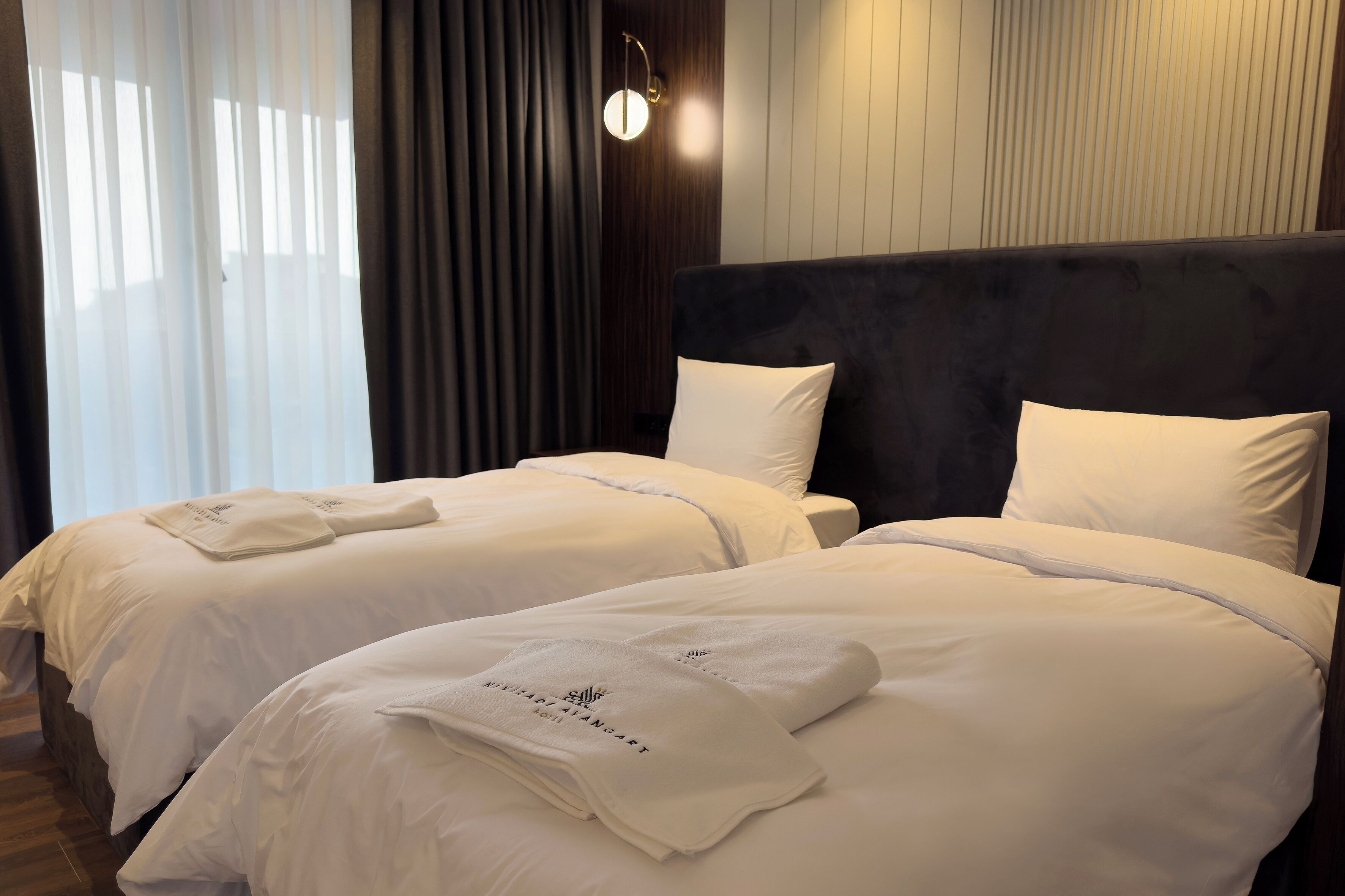 A hotel room with two beds, each with white linens and towels, a dark headboard, and a wall-mounted light fixture.