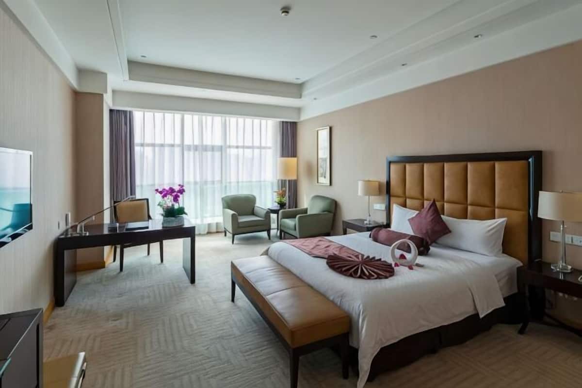 A hotel room with a large bed, a desk, two chairs, a television, and a window with curtains.
