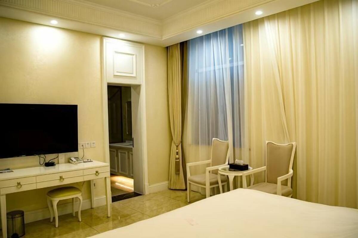 A hotel room with a bed, a desk with a television, two chairs, and a window with curtains.