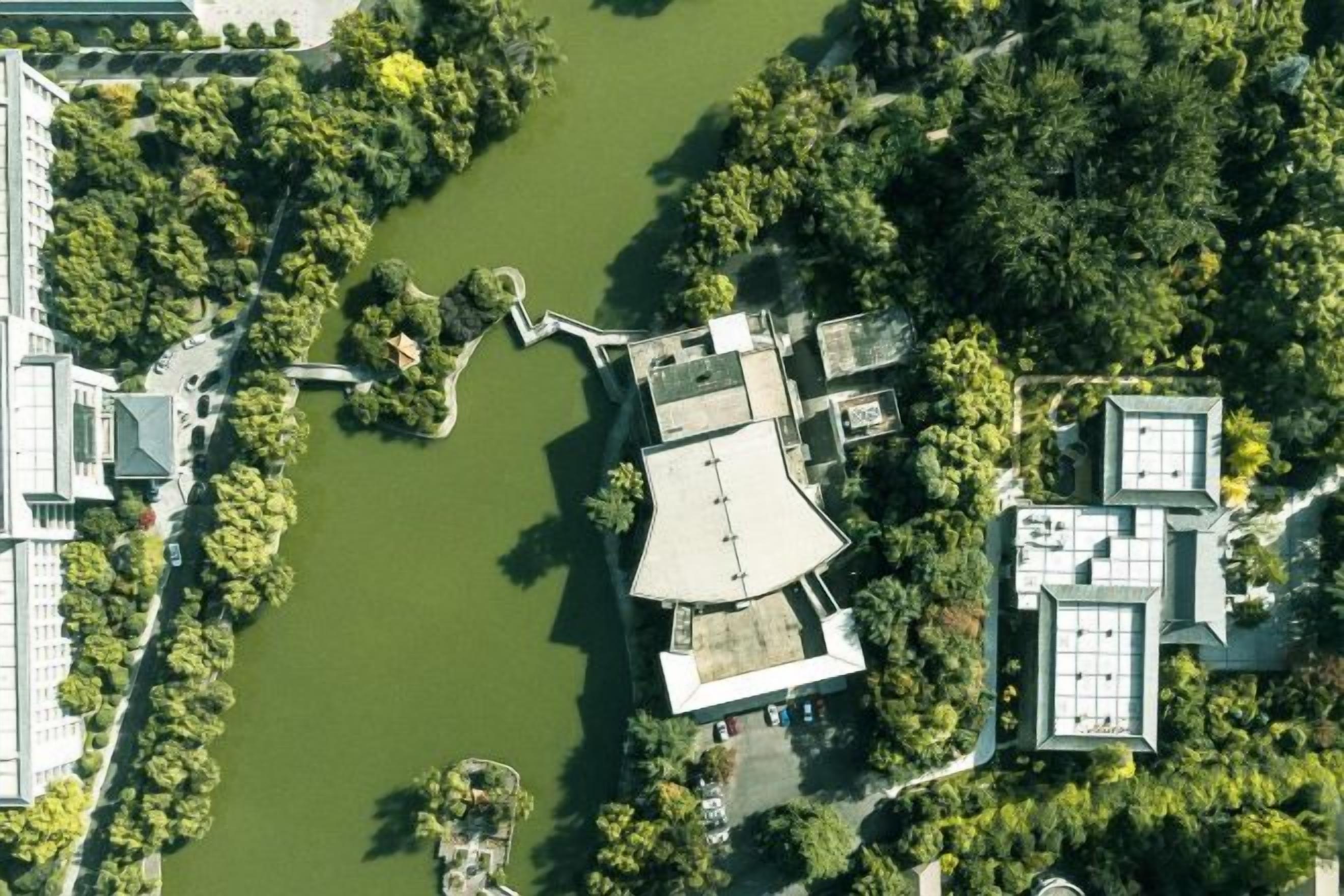 An aerial view of a building complex with a central courtyard, surrounded by dense greenery and a body of water.