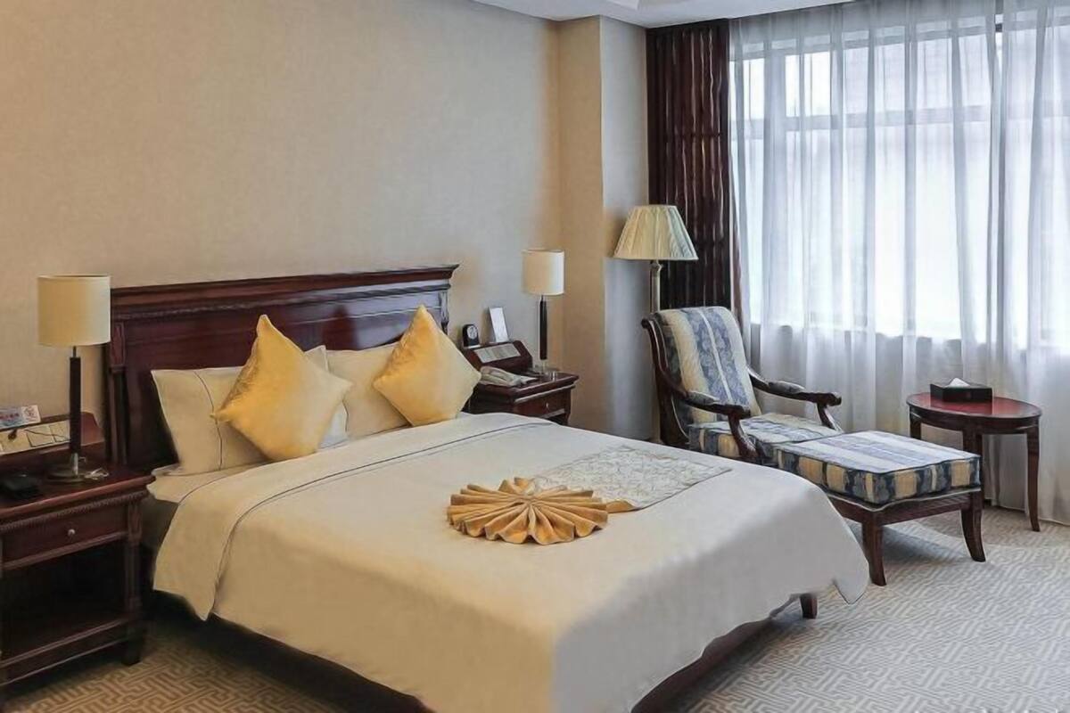 A hotel room with a large bed, two bedside tables, a chair, a lamp, and a view of the outdoors through sheer curtains.