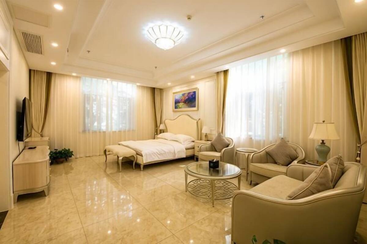 A well-lit, spacious hotel room with a bed, two armchairs, a sofa, a coffee table, and a TV.