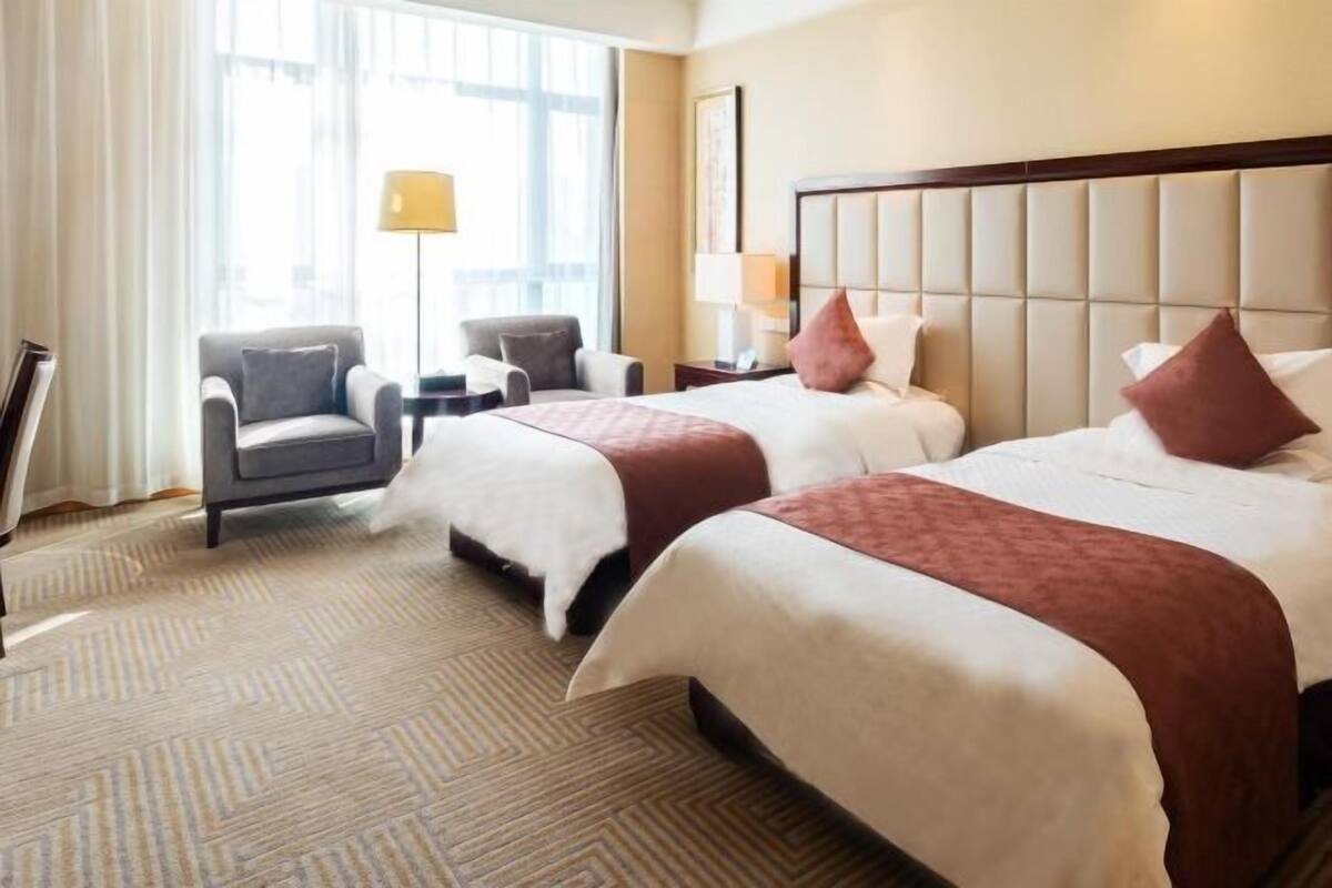 A hotel room with two beds, a sitting area with a chair and a small table, and a large window providing natural light.