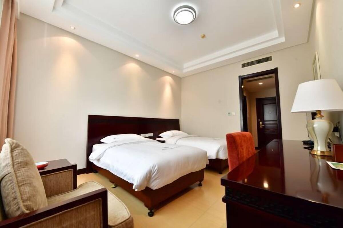 A hotel room with two beds, a desk, a chair, a lamp, and a door leading to another room.