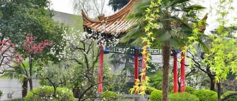A traditional pavilion with a curved roof, surrounded by lush greenery and flowering trees.