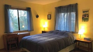 A bedroom with a bed, two bedside tables with lamps, a chair, and two windows with curtains.