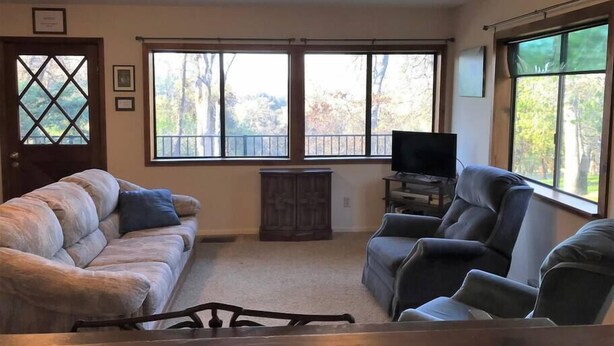 A living room with a beige sofa, a dark armchair, and a TV stand. There are two large windows with a view of trees and a balcony.