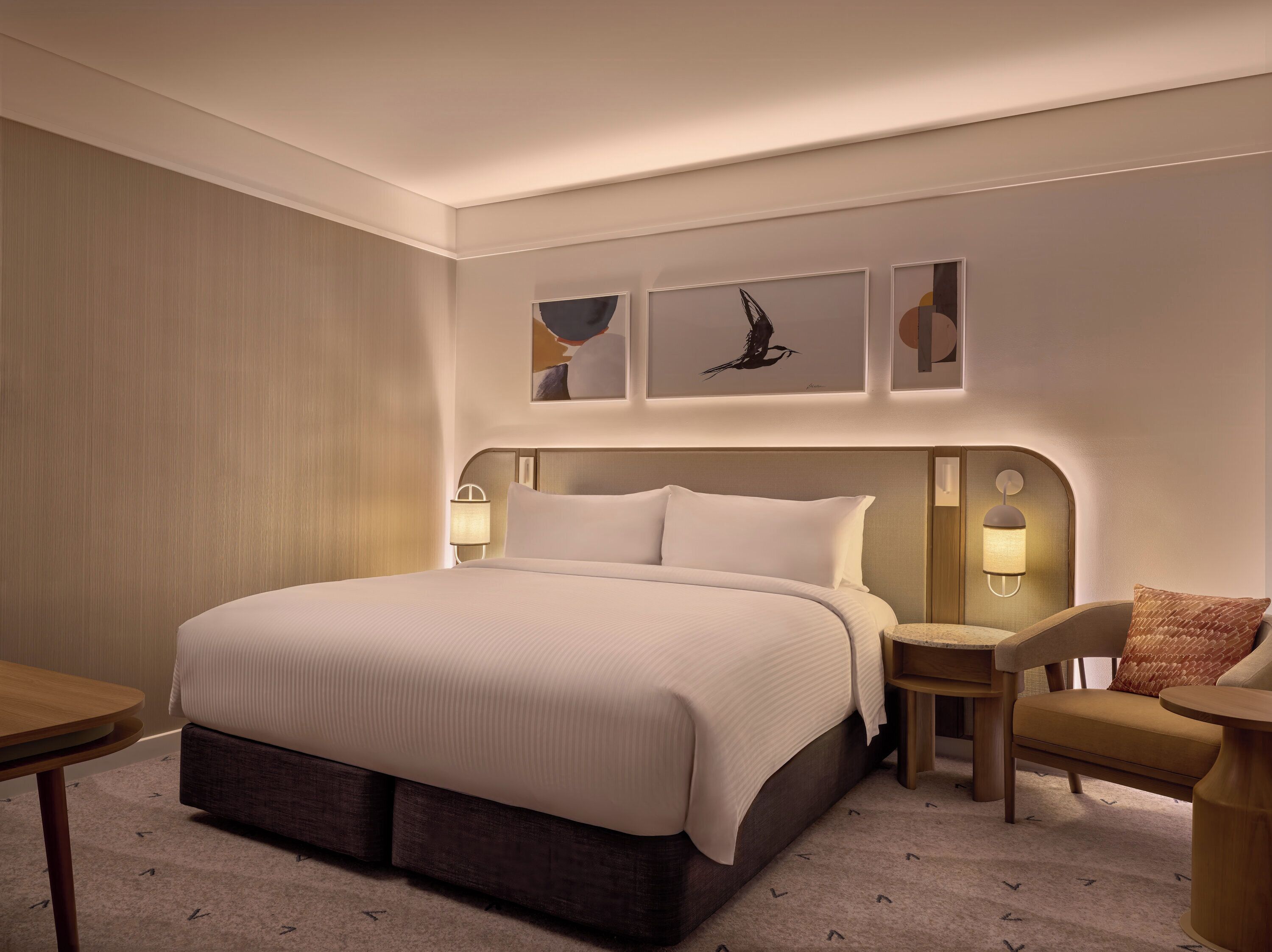 A modern hotel room with a large bed, bedside lamps, a small table, a chair, and artwork on the wall.