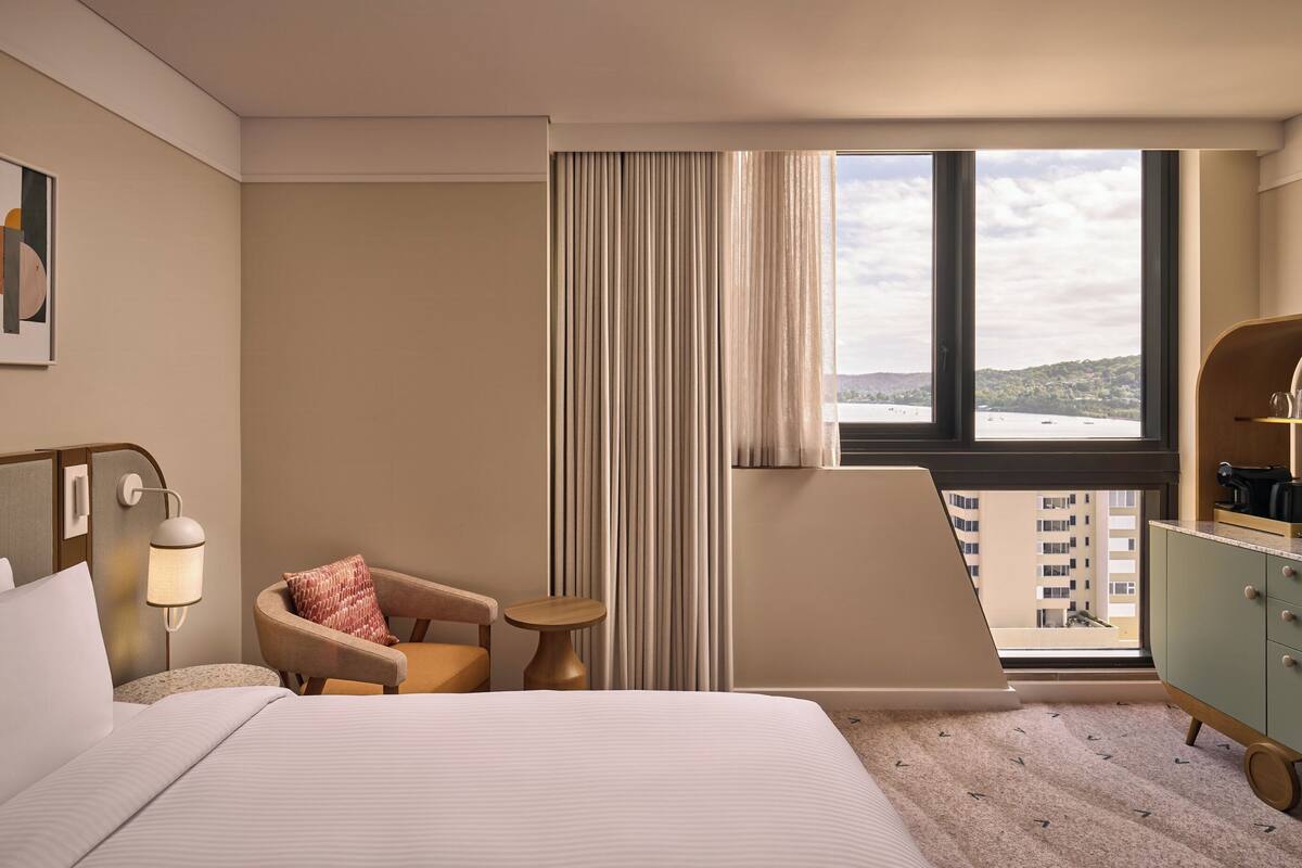A hotel room with a bed, a chair, a small table, a lamp, a cabinet, and a window with a view of a body of water and buildings.