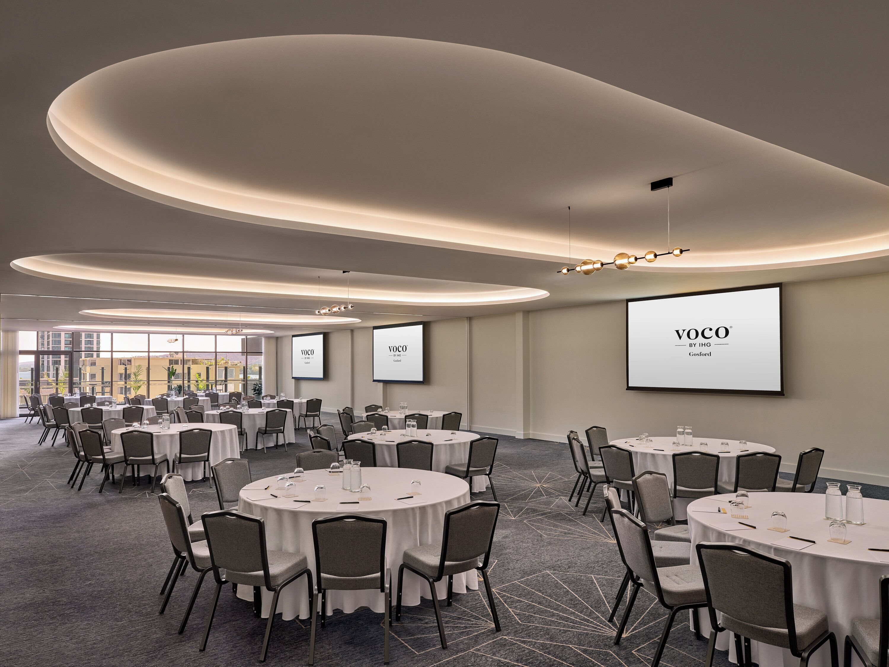 Un salón de conferencias con varias mesas redondas, sillas y pantallas grandes que muestran el logotipo de "VOCO".