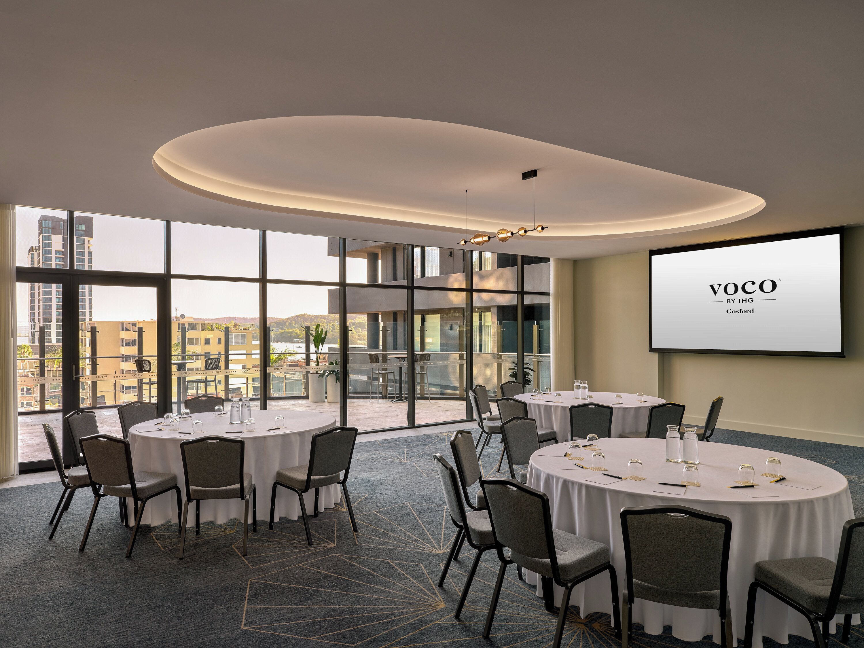 Ein Konferenzraum mit runden Tischen, Stühlen und einem großen Bildschirm, der "VOCO BY IHG Gosford" anzeigt.
