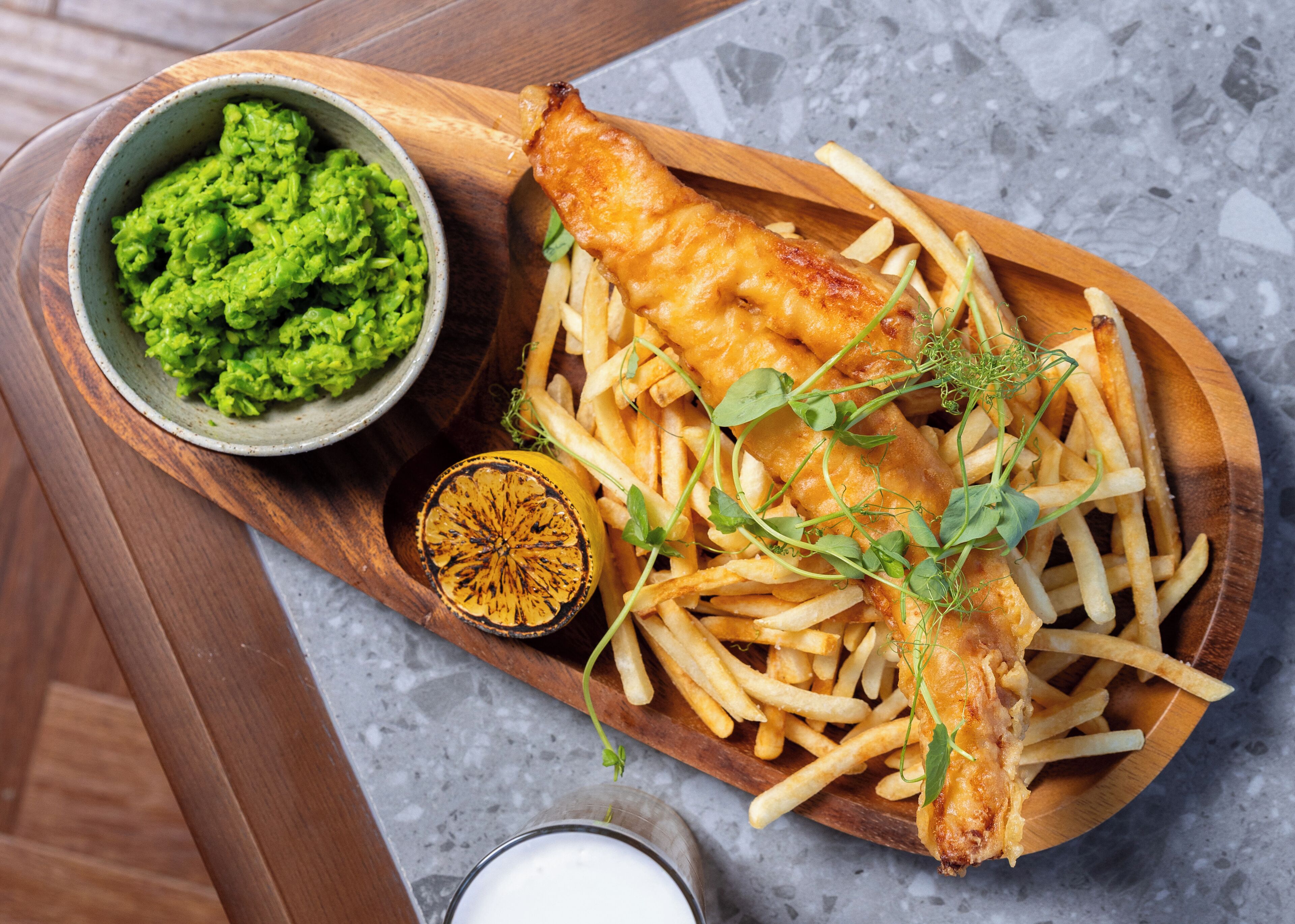 A wooden serving board with fish and chips, a bowl of mushy peas, and a lemon half.