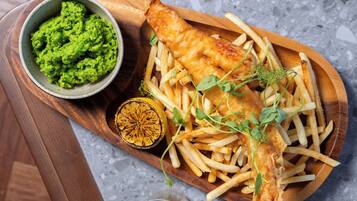 A wooden serving board with fish and chips, a bowl of mushy peas, and a lemon half.