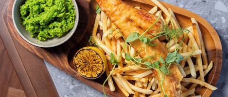 A wooden serving board with fish and chips, a bowl of mushy peas, and a lemon half.