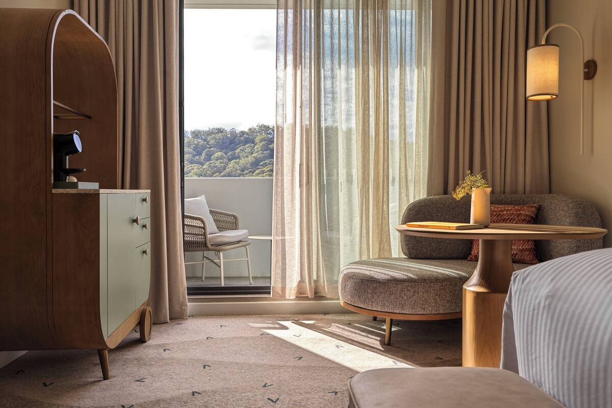 A modern hotel room with a curved wooden cabinet, a round wooden table, a comfortable armchair, and a sofa.
