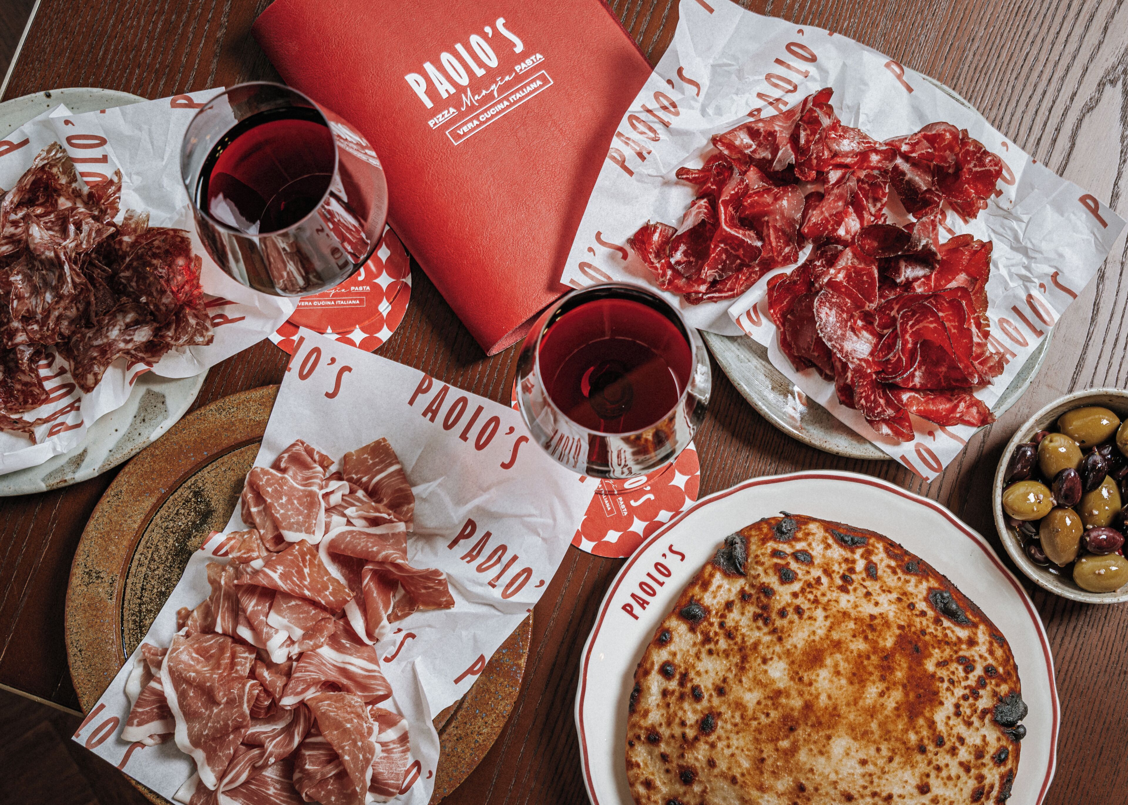 Una mesa con diversos alimentos, como platos de embutidos, una pizza y un bol de aceitunas.