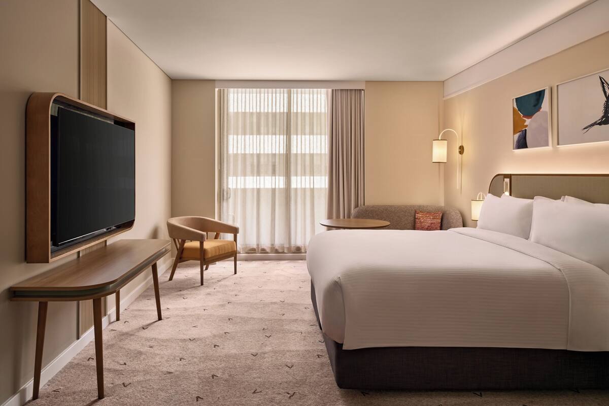 A hotel room with a large bed, a TV mounted on a wooden console, a small table, a chair, and a window with curtains.