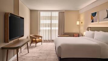 Habitación de hotel con una cama grande, un televisor montado en una consola de madera, una mesita, una silla y una ventana con cortinas.
