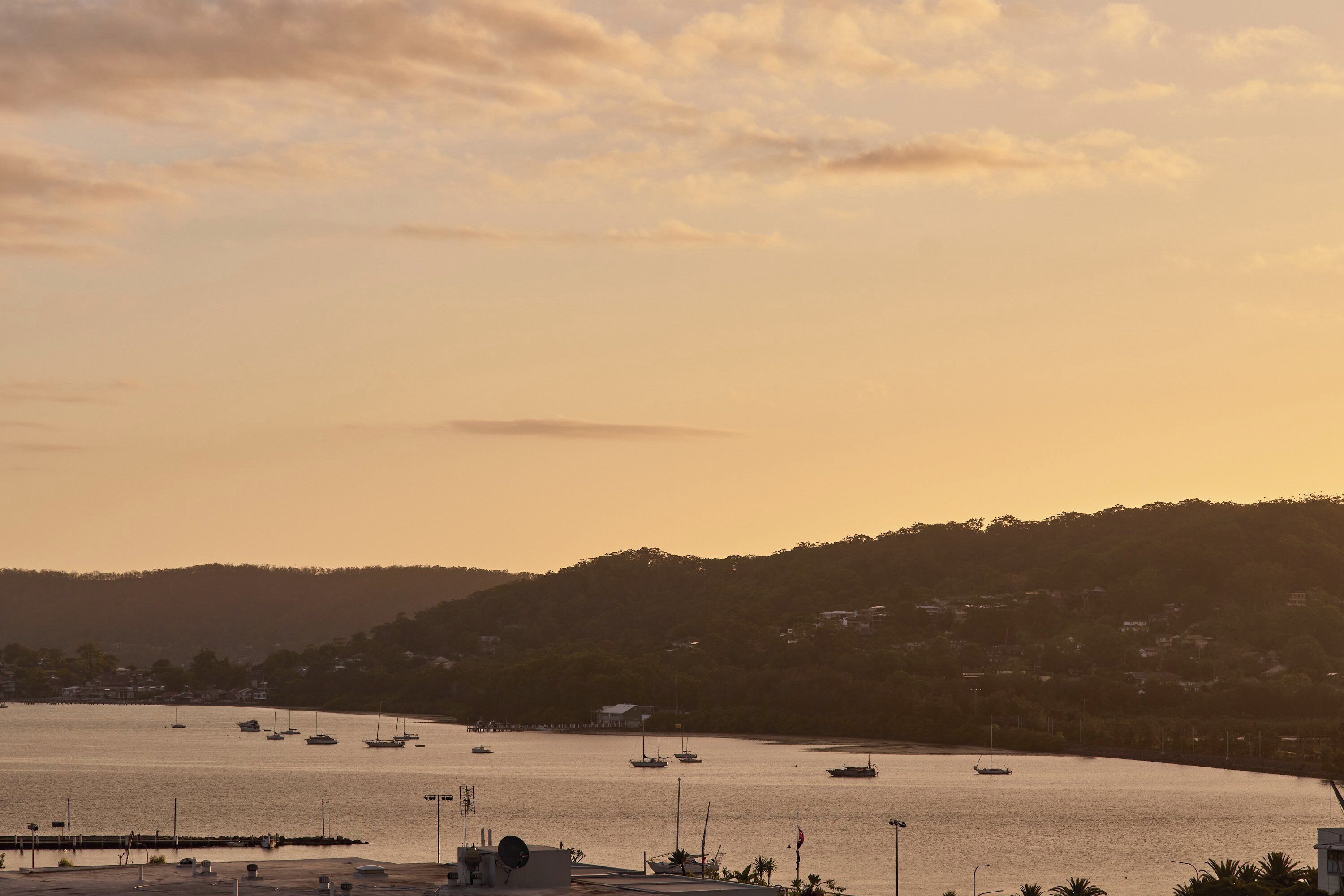 Vista costera con barcos amarrados en un puerto deportivo, un muelle y un paisaje montañoso al fondo.