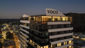 Un edificio moderno con un letrero que dice "VOCO", con varios pisos y grandes ventanales, ubicado en una zona urbana con calles y edificios visibles.