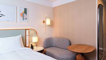 A hotel room with a bed, a gray armchair, a small round table, and a wall-mounted lamp.