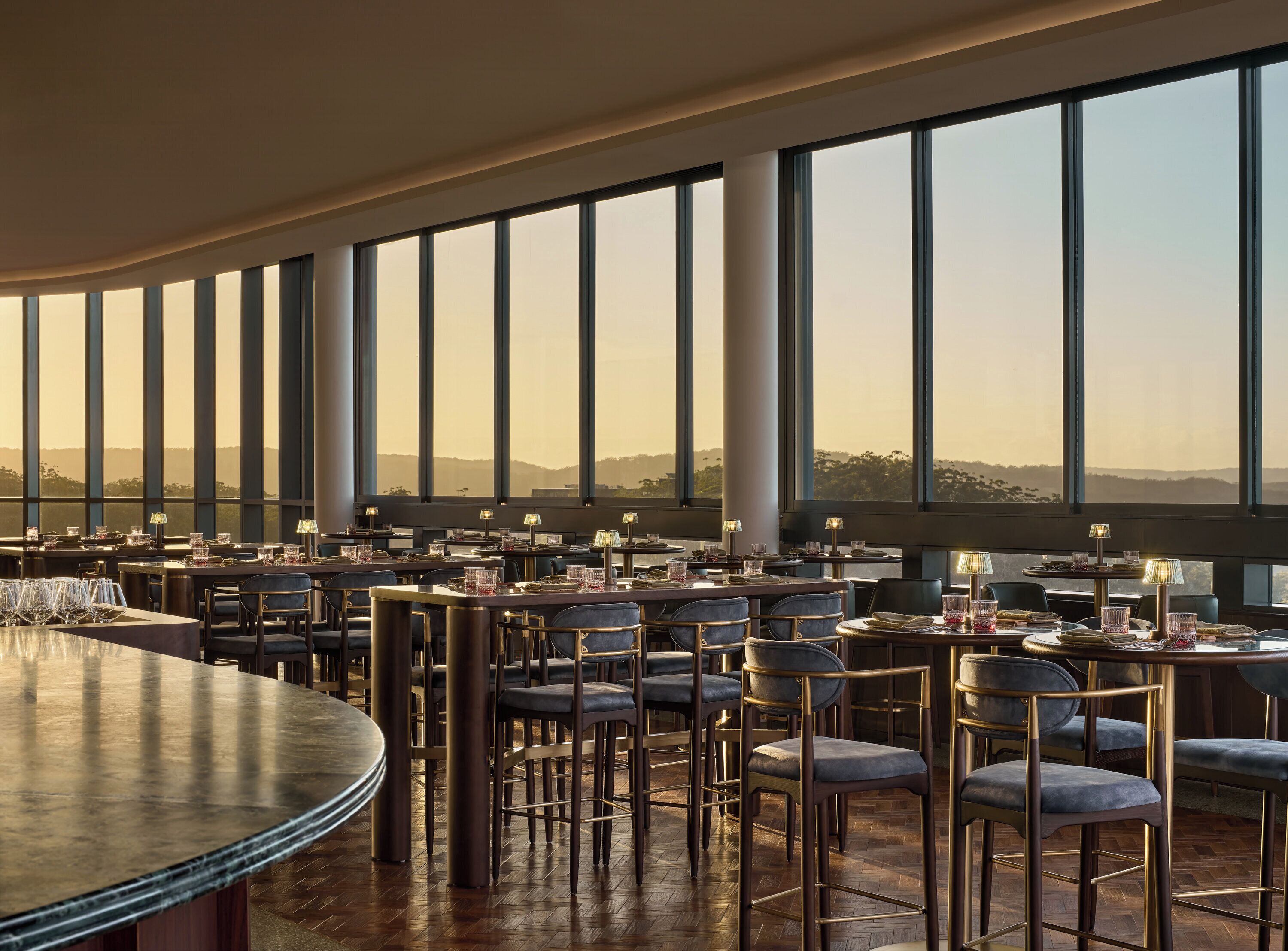 A restaurant with high ceilings, large windows, and a view of the hills. Tables are set with glasses and cutlery.