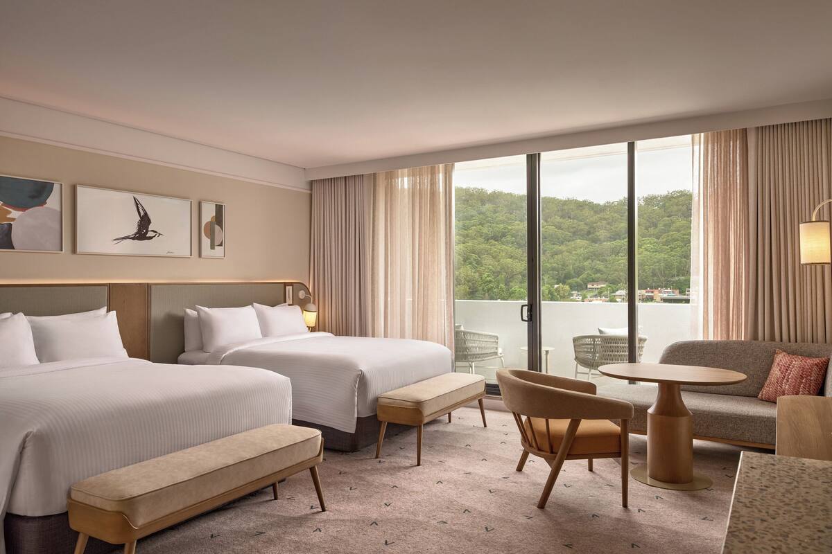 A hotel room with two beds, a sofa, a table, and a balcony with a view of greenery.