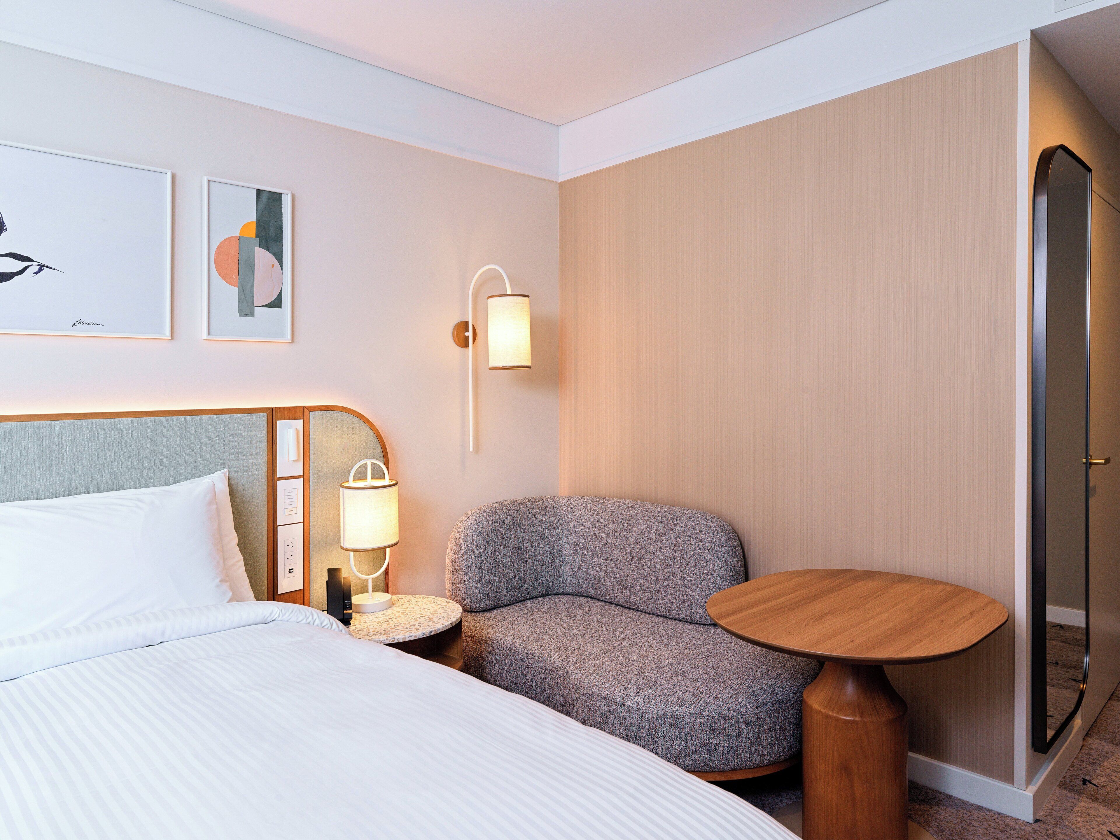 Habitación de hotel con cama, un sillón gris, una mesita redonda, una lámpara de pared y una puerta.