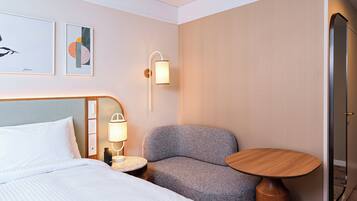 Habitación de hotel con cama, un sillón gris, una mesita redonda, una lámpara de pared y una puerta.