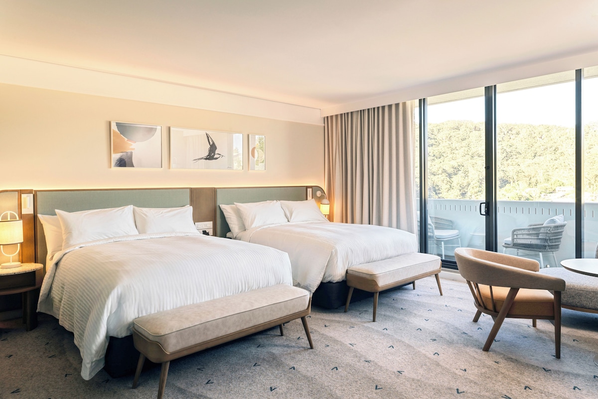 Habitación de hotel con dos camas, un banco, una silla y un balcón con mobiliario exterior.