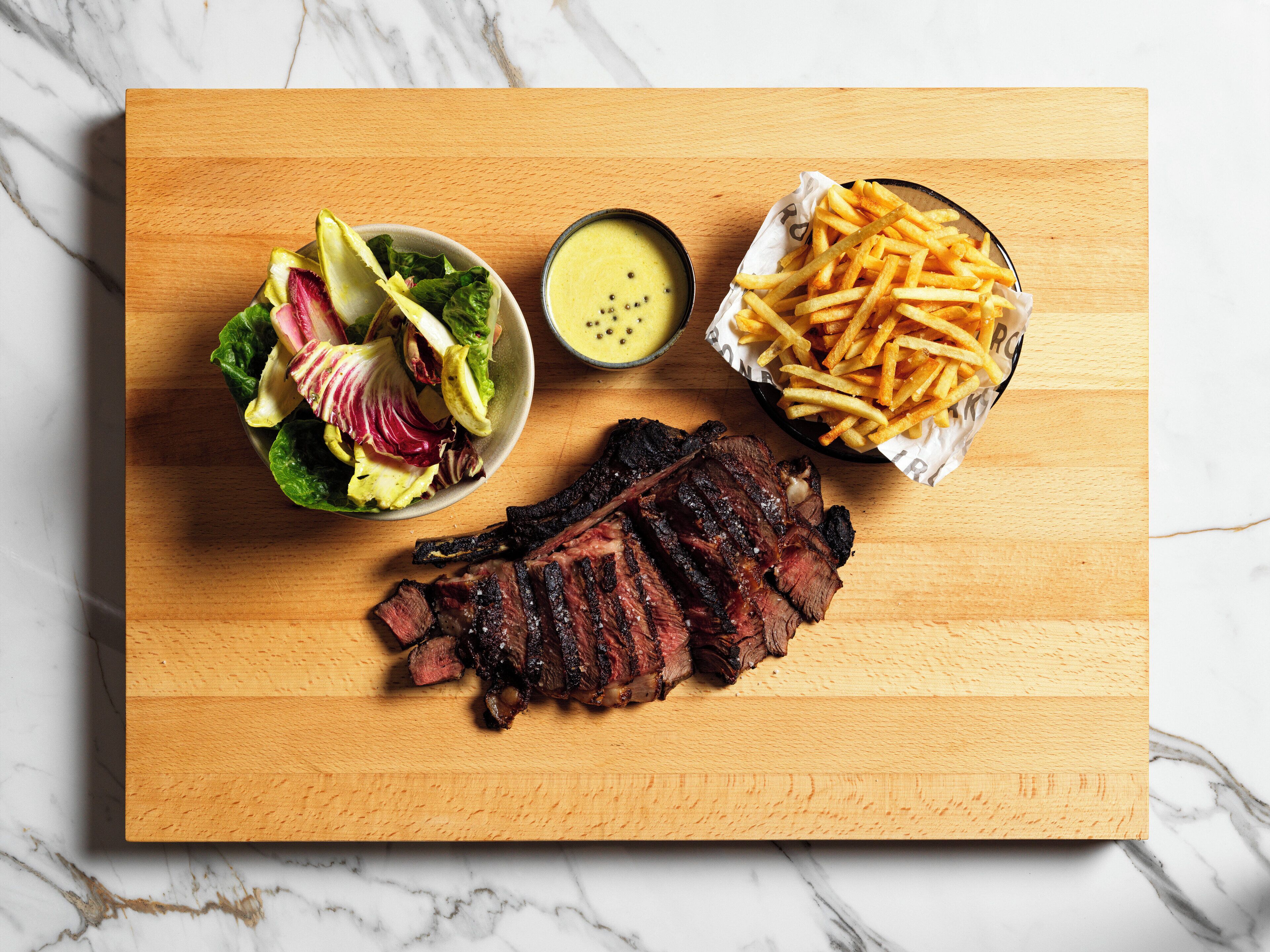 Una tabla de madera con un bistec a la parrilla, un tazón de ensalada, un recipiente pequeño de salsa y una cesta de papas fritas.