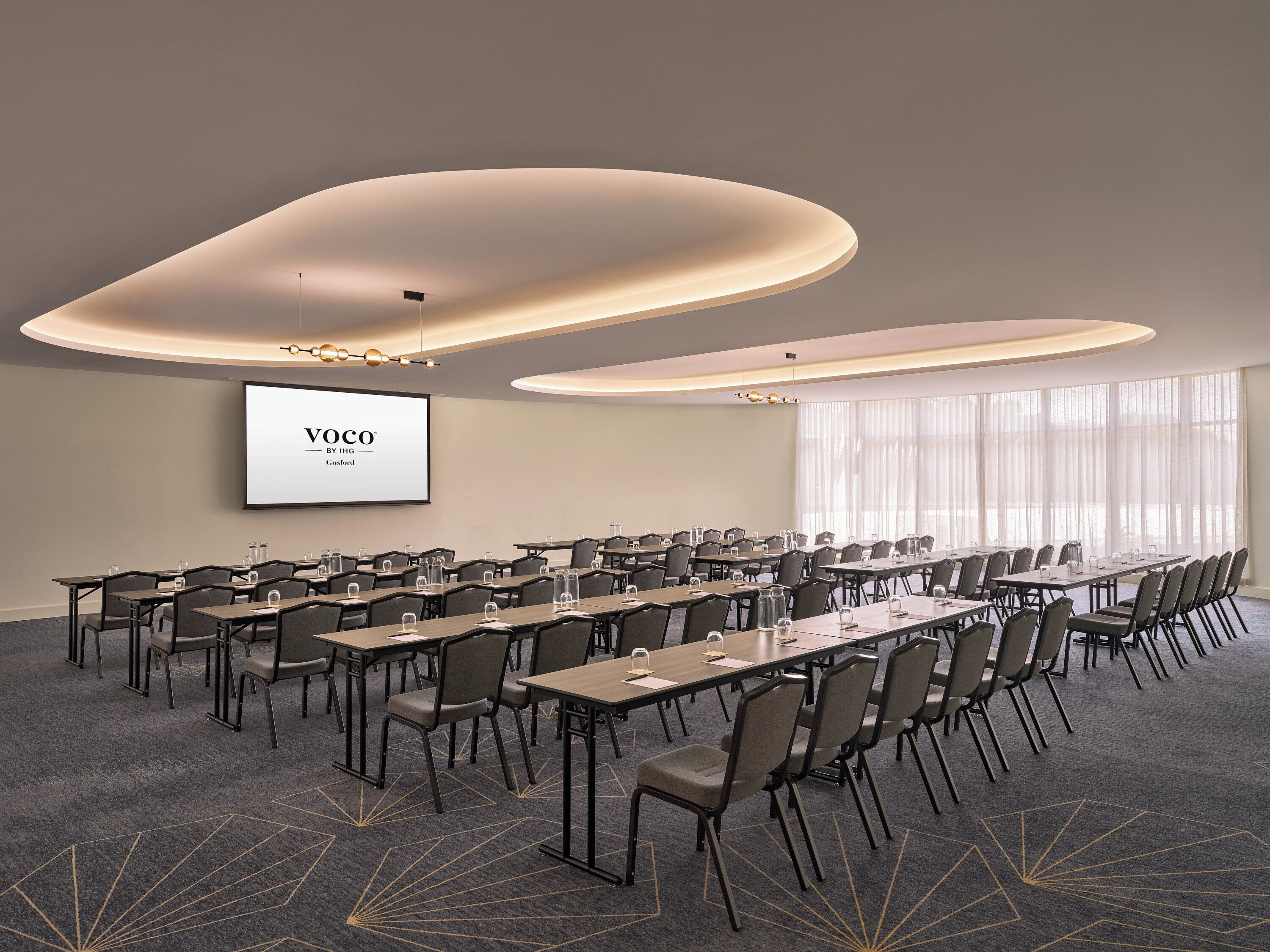 A conference room with multiple tables and chairs, a large screen displaying "VOCO BY IHG Gosford", and a modern ceiling design.